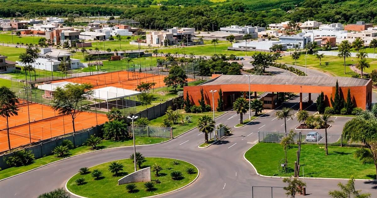 Vista aérea da portaria de tijolos aparentes, com rotatória, quadras de tênis de saibro e casas ao fundo, cercadas por vegetação.