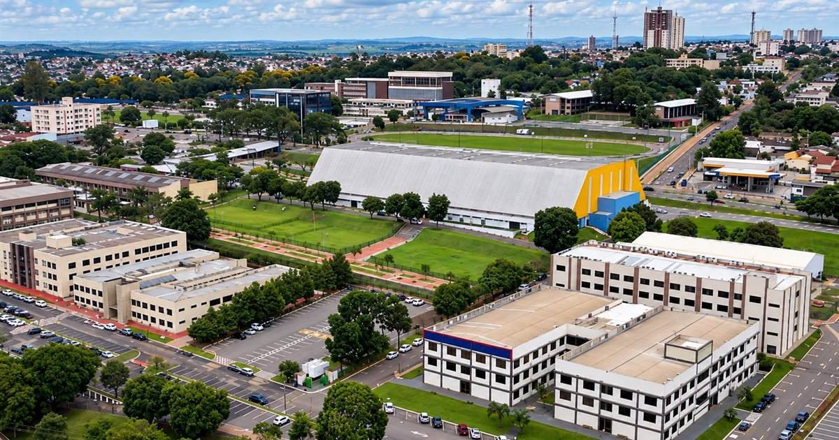 Vista aérea de um campus com prédios de salas, ginásio coberto, campo e estacionamentos, com a cidade ao fundo.
