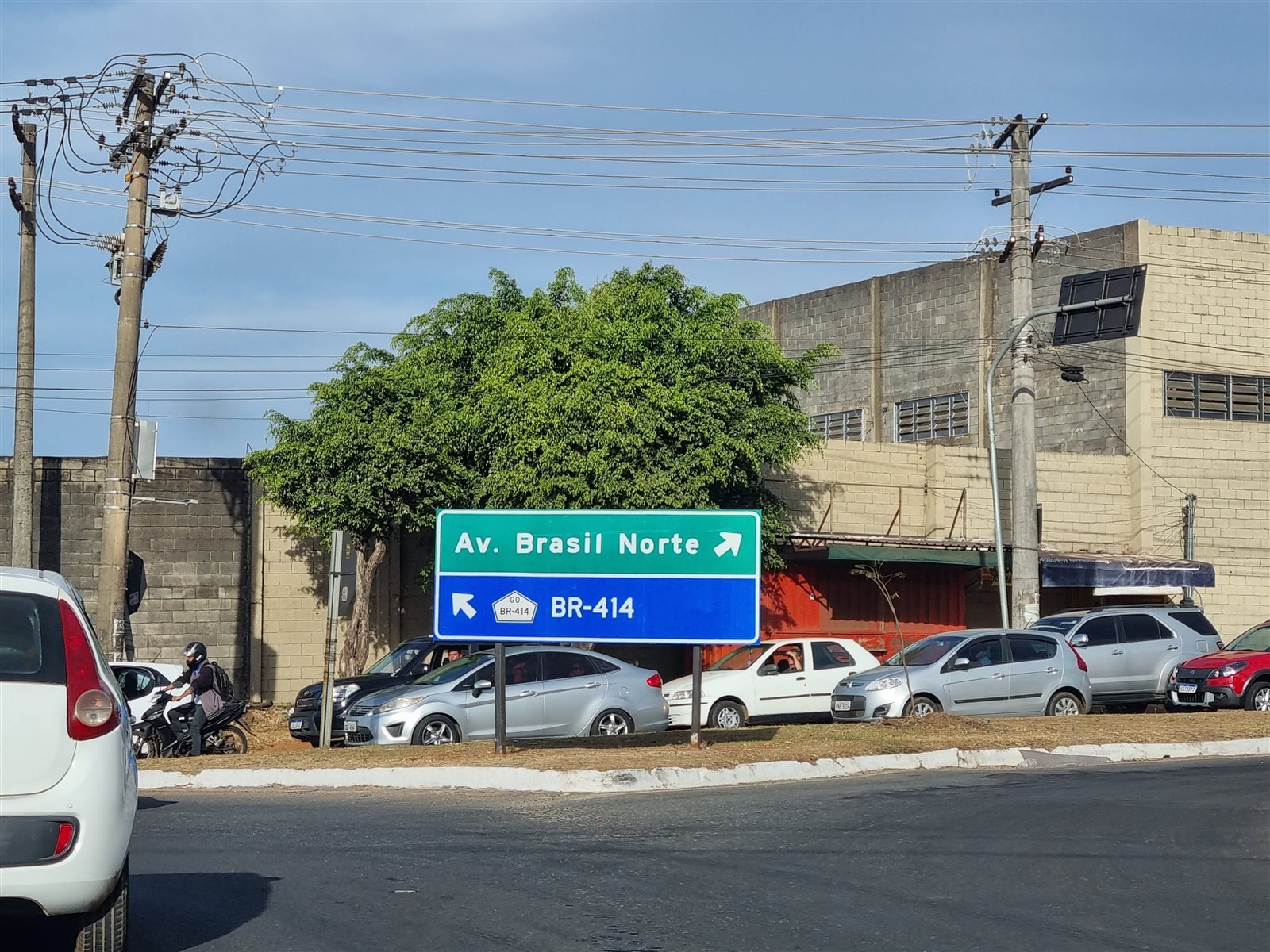 Placa indicando Avenida Brasil Norte e BR-414 em um cruzamento, com galpões ao fundo.