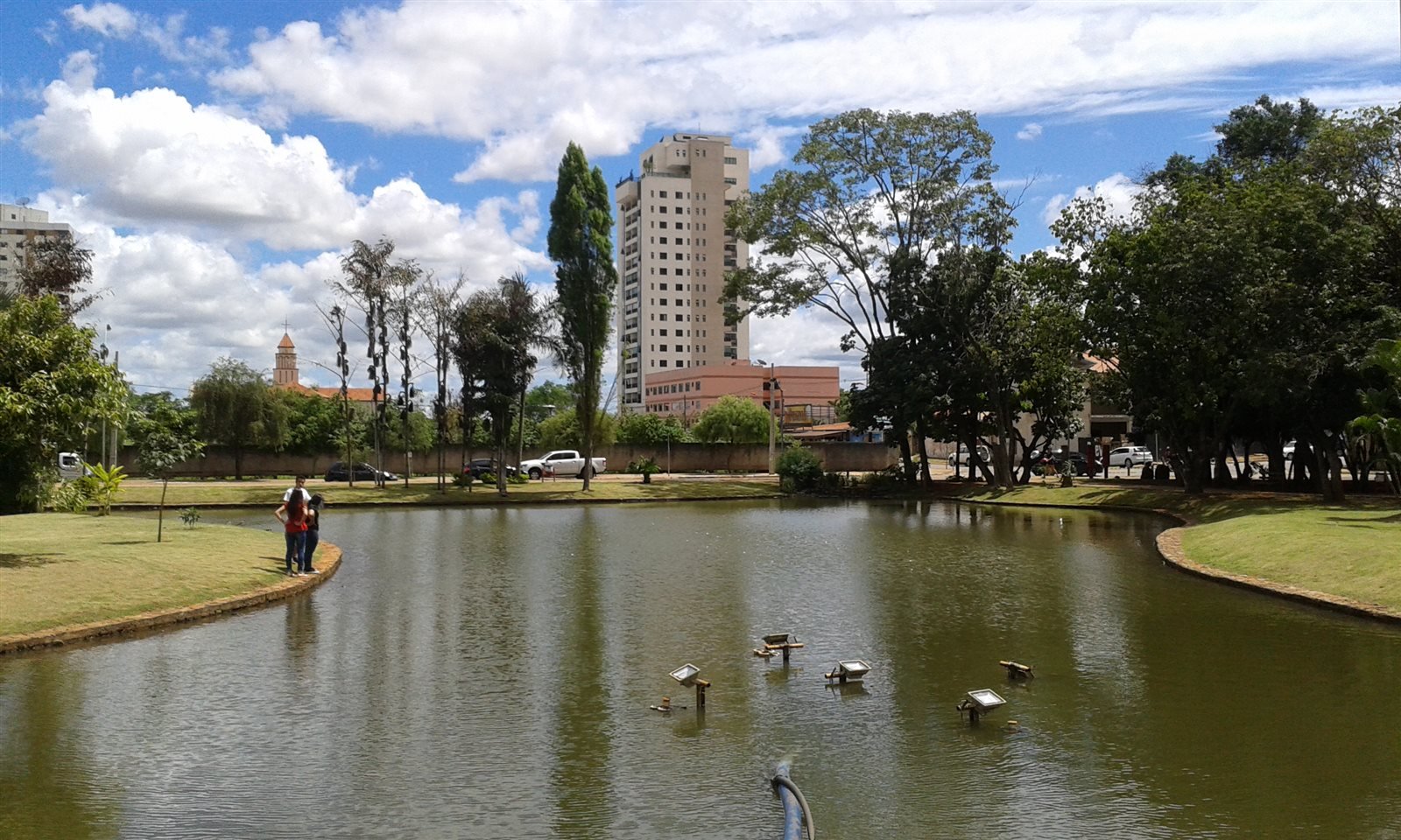 Lago cercado de gramado e árvores, com um edifício residencial alto ao fundo.